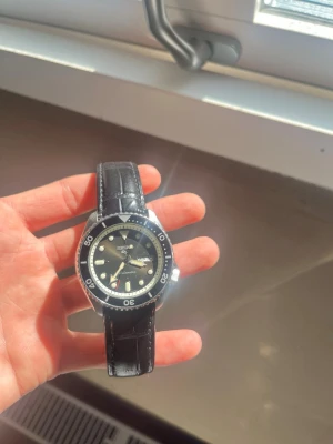 Svart Seiko automatisk klocka - Stilren Seiko automatisk klocka med svart urtavla och tydliga index. Klockan har en klassisk rund form och svart läderarmband med krokodilpräglat mönster. Datum- och veckovisning på urtavlan. Perfekt för dig som gillar tidlös design.
