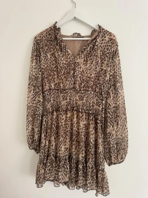 Leopardmönstrad kortklänning - Kort klänning med leopardmönster i beige och bruna toner. Klänningen har v-ringning, långärmade puffärmar och midjan ger en snygg passform. Volangdetalj nedtill ger extra rörelse och stil.
