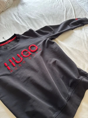 Svart sweatshirt från Hugo Boss - Svart sweatshirt från Hugo Boss med röd broderad HUGO-logga på bröstet och ärmen. Tröjan har rund halsringning, långa ärmar och ribbade muddar. Perfekt för en stilren och trendig look.