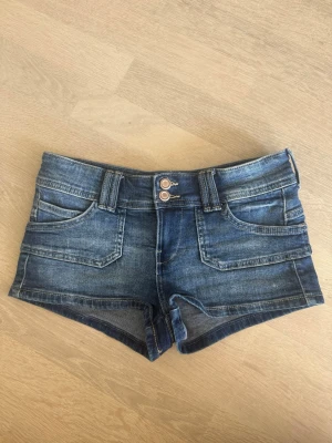 Blå jeansshorts med broderade kronor - Snygga blå jeansshorts med dubbla knappar framtill och broderade rosa kronor på bakfickorna. Klassisk femficksmodell med bälteshällor och korta ben. Perfekta för varma dagar och en avslappnad stil. Pris kan diskuteras!