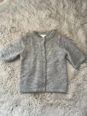 Grå kofta från Gina Tricot - Mysig grå kofta från Gina Tricot med rund halsringning och pärlknappar framtill. Koftan har halvlånga ärmar och är stickad i mjukt material. Perfekt att slänga över en topp för en chill look. Knappt använd och passar jätte bra över ett vitt linne 🫶🏻