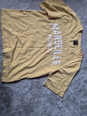 Gul t-shirt med Marseille-tryck - Snygg gul t-shirt med texten 'MARSEILLE FRANCE' i vitt framtill. Klassisk rund halsringning och korta ärmar. Perfekt för dig som gillar streetstyle och vill ha en chill vibe i garderoben.