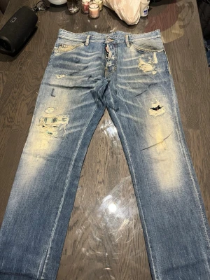 Blå slitna jeans från Dsquared2 - Säljer ett par blå jeans från Dsquared2 med distressed detaljer och slitningar på både fram- och baksida. Jeansen har klassisk femficksmodell, knappgylf och en snygg tvättad look. Perfekta för dig som gillar en edgy och trendig stil.
