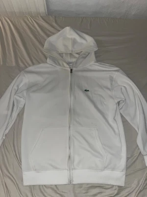 Vit hoodie med dragkedja från Lacoste - Snygg vit hoodie från Lacoste med dragkedja framtill och klassisk krokodillogga på bröstet. Hoodien har huva, två fickor framtill och ribbade muddar. Perfekt för en clean och stilren look.