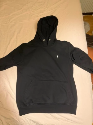 Svart hoodie från Polo Ralph Lauren - Säljer en svart hoodie från Polo Ralph Lauren med klassisk vit logga på bröstet. Hoodien har stor huva med metallöljetter och en rymlig magficka framtill. Perfekt för en avslappnad och stilren look.