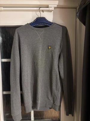 Grå sweatshirt med broderad logga - En stilren grå sweatshirt med rund hals och lång ärm. Tröjan har en liten broderad gul logga på bröstet och ribbade muddar vid ärmslut och nederkant. Perfekt för en avslappnad och trendig look.