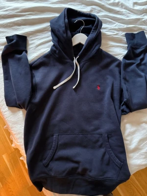 Mörkblå hoodie från Polo Ralph Lauren - Snygg mörkblå hoodie från Polo Ralph Lauren med klassisk huva och vita dragsnören. Har en känguruficka framtill och den ikoniska röda loggan broderad på bröstet.🤩 OBS, tröjan har en liten skavank (se sista bilden) på slutet av ärmen, syns dock knappt så är ingen större fara.