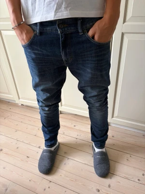 Mörkblå East West slim fit jeans herr - Säljer ett par mörkblå slim fit jeans med snygga slitningar och klassisk femficksmodell. Jeansen har normal midja och smal passform genom benen. Perfekta för en avslappnad men ändå stilren look. Jeansen har en liten fläck på höger bakficka men borde gå bort i tvätten. Jeansen har storlek 32/32. Hör av er vid frågor och priset är inte hugget i sten!