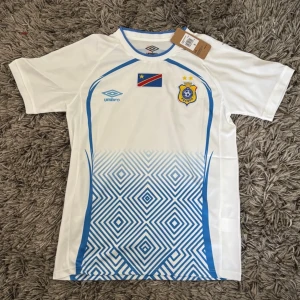 Vit fotbollströja Umbro ECOFF - Snygg vit fotbollströja från Umbro med blå detaljer och geometriskt mönster nedtill. Tröjan har ECOFF-emblem och flagga på bröstet samt rund halsringning med blå kant. Perfekt för träning eller match.