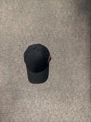 Svart keps med mönster och röd detalj - Säljer en svart keps med diskret mönster och en röd detalj på sidan. Kepsen har böjd skärm och justerbart spänne bak för perfekt passform. Materialet ser ut att vara bomull eller canvas, vilket gör den både snygg och bekväm.