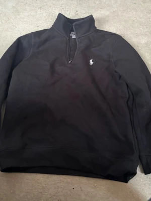Svart halvzip tröja från Polo Ralph Lauren - Klassisk svart halvzip tröja från Polo Ralph Lauren med den ikoniska vita polospelaren broderad på bröstet. Tröjan har hög krage och långa ärmar, perfekt för en stilren och avslappnad look.