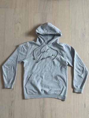 Saint Laurent hoodie - Snygg grå Saint Laurent hoodie med huva och magficka. Perfekt för en avslappnad och trendig look. En sällsynt modell som man inte ser ofta i storlek S, vid fler frågor är det bara att höra av sig!
