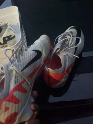 Nike vita och röda fotbollsskor - Säljer ett par Nike fotbollsskor i vitt med röda och svarta detaljer. Skorna har snörning och är designade för bra grepp på planen. Snygg och modern look med stora loggor på sidorna. Perfekta för dig som vill sticka ut på fotbollsplanen.