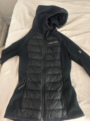 Svart pufferjacka från Peak Performance - Snygg svart pufferjacka från Peak Performance med huva och dragkedja framtill. Jackan har quiltade partier på framsidan och släta ärmar, samt två dragkedjeförsedda fickor. Perfekt för kalla vinterdagar och riktigt stilren design. Köpte denna för 3000 men säljer den för 1200 