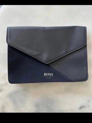 Mörkblå necessär från Hugo Boss - Stilren mörkblå necessär / kuvertväska från Hugo Boss med elegant triangulärt lock och diskret logga framtill. Den ena delen är i mockaliknande tyg och resten är i polyuretan. Väskan har en minimalistisk design som passar perfekt till alla tillfällen. 
