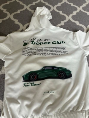 Vit hoodie med biltryck och text - Säljer en vit hoodie med huva och känguruficka. På ryggen finns ett stort tryck med en grön sportbil och texten 'Champagne St Tropez Club' samt '911 Club From Monaco'. Framtill finns en liten logga med text. Perfekt för dig som gillar bilar och streetwear.