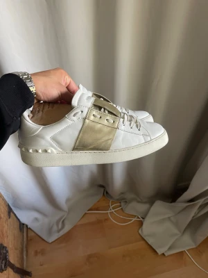 Valentino Open  - Säljer ett par vita sneakers från Valentino med en bred guldfärgad detalj över snörningen och nitar på hälen. Skorna är låga och tillverkade i läder med klassisk siluett och vita snören. Perfekta för dig som vill ha något stilrent men ändå sticker ut.