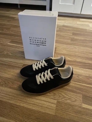Maison Margiela svarta sneakers - Svarta sneakers från Maison Martin Margiela med vita snören och beige innersula. Skorna har en klassisk låg siluett och är tillverkade i en mix av mocka och läder. Snygg gummisula och stilren design som passar till det mesta.