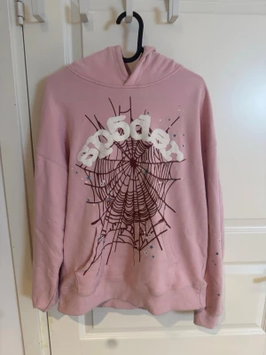 Rosa hoodie med spindelnätsmotiv - Cool rosa hoodie med stor spindelnätsgrafik och texten 'sp5der' i vitt på bröstet. Hoodien har huva och ribbade muddar vid ärmslut och nederkant. Materialet är mjukt och bekvämt, perfekt för en avslappnad streetwear-look.