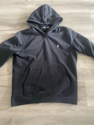 Svart hoodie från Polo Ralph Lauren - Snygg svart hoodie från Polo Ralph Lauren med klassisk vit broderad logga på bröstet. Hoodien har en stor känguruficka framtill och en skön huva. Tillverkad i mjukt bomullsmaterial som är perfekt för chill dagar.
