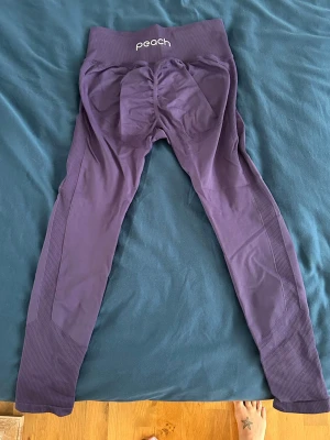 Lila seamless leggings från Peach - Säljer ett par lila seamless leggings från Peach med hög midja och ribbade detaljer. Materialet är stretchigt och följsamt, perfekt för träning eller chill. Diskret logga på ena benet och scrunch-detalj bak för extra form. Använda men i fint skick. Storlek S.