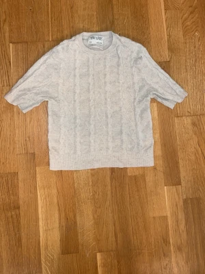 Beige kabelstickad tröja AWARE - Beige kabelstickad tröja från AWARE med korta ärmar och rund hals. Tröjan har ett diskret mönster framtill och är gjord i ett mjukt stickat material. Perfekt för lager på lager och enkel att matcha till olika outfits.