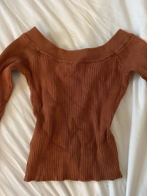 Rostbrun ribbad offshoulder topp - Säljer en snygg ribbad offshoulder topp i en varm rostbrun färg. Toppen har lång ärm och är figurnära med stretchigt material. Perfekt att styla med jeans eller kjol för en trendig look.