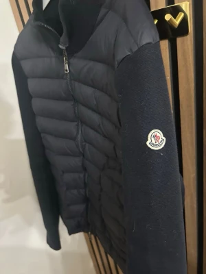 Mörkblå cardiganjacka från Moncler - Snygg mörkblå cardiganjacka från Moncler med quiltad framsida och stickade ärmar. Dragkedja framtill och Moncler-logga på ärmen. Perfekt för dig som vill ha en stilren och exklusiv look. Materialmix av dun och ull ger både värme och komfort. Den har ett litet håll, som ni ser på bilden som inte syns.
