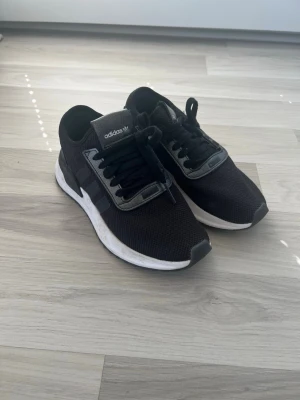 Svarta Adidas sneakers med mesh - Snygga svarta Adidas sneakers med ovandel i mesh och detaljer i grått. Skorna har klassiska tre ränder på sidan, svart snörning och vit sula. Perfekta för en sportig och stilren look. 
