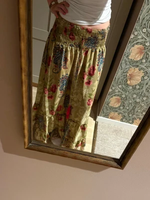 Blommig långkjol med paisleymönster - Säljer en flowy långkjol i beige med blommigt och paisleyinspirerat mönster i rött, blått och grönt. Kjolen har bred smock i midjan och volang längst ner. Perfekt för dig som gillar boho-vibbar och färgglada prints.