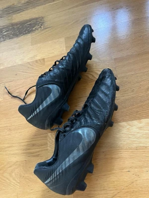 Svarta Nike fotbollsskor i läder - Säljer ett par svarta Nike fotbollsskor med klassisk snörning och dobbar för spel på gräs. Skorna har en stilren design med stora silvriga Nike-loggor på sidorna och är tillverkade i mjukt läder för bra bollkontroll och komfort.