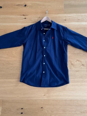 Mörkblå skjorta från Polo Ralph Lauren - Klassisk mörkblå skjorta från Polo Ralph Lauren med vit knappstängning och röd broderad logga på bröstet. Skjortan har lång ärm, normal passform och är tillverkad i bomull. Perfekt för en clean och stilren look.