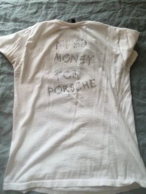 Vit t-shirt med strasstext från BVALDI - Vit t-shirt med korta ärmar och rund hals. På bröstet finns initialerna 'NB' i strass och på ryggen står det 'NEED MONEY FOR PORSCHE' i glittriga stenar. Perfekt för dig som gillar statement-plagg och vill sticka ut lite extra. BVALDI 