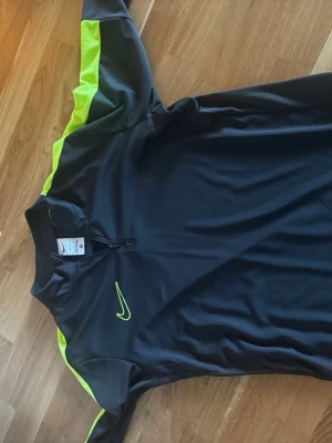 Nike svart träningsjacka med neon - Snygg svart träningsjacka från Nike med neonfärgade detaljer på axlar och ärmar. Jackan har halv dragkedja framtill och är tillverkad i ett lätt, sportigt material som passar perfekt till träning eller uppvärmning. M storlek men sitter rätt så tajt.