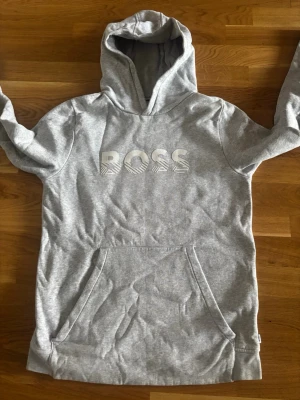 Grå hoodie från BOSS med huva - Snygg grå hoodie från BOSS med stor huva och magficka. Trycket BOSS i vitt framtill ger en clean look. Tillverkad i mjukt och bekvämt material, perfekt för chill dagar. Klassisk passform och ribbade muddar vid ärmar och nederkant. Ser lite skrynklig ut på bilder men det är pga kamera. Ser inte ut så i verkligheten 