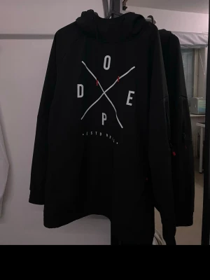 Svart jackhoodie från Dope med tryck - Svart jacka som ser ut som en hoodie från Dope med stort vitt tryck på bröstet där bokstäverna D-O-P-E är placerade i ett kors. Hoodien har huva med dragsko och är fodrad med mesh på insidan för extra komfort. Perfekt för en sportig och avslappnad stil. Storlek M