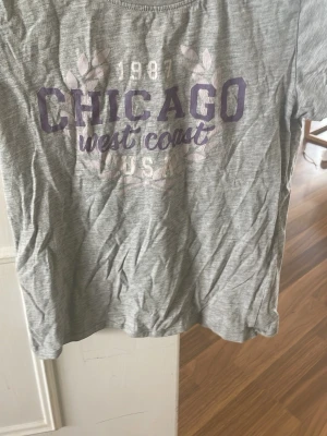 Grå t-shirt med CHICAGO-tryck - Snygg grå t-shirt med lila och vitt tryck där det står 'CHICAGO west coast USA 1987' framtill. Klassisk rund halsringning och korta ärmar. Perfekt för en avslappnad och cool stil.