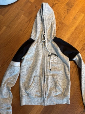 Grå zip hoodie från Hollister - Snygg gråmelerad hoodie från Hollister med dragkedja, huva med vita snören och svarta kontrastärmar. Broderad text och siffran 22 på bröstet. Två stora fickor framtill och ribbade muddar. Perfekt för en chill och sportig look.