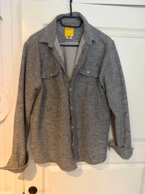 Grå skjorta med knappar/ overshirt  - Säljer en stilren grå skjorta/overshirt med knappar framtill och två bröstfickor. Skjortan har lång ärm och klassisk krage, perfekt för en avslappnad men snygg look. Materialet känns mjukt och bekvämt mot huden. Storlek M köparen står för frakt 