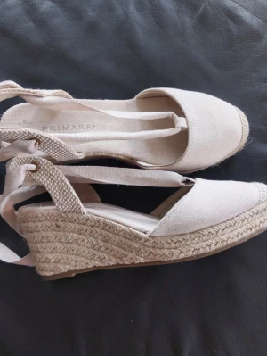Beiga kilklack sandalet från Primark - Snygga beiga sandaler med kilklack och med knytband runt ankeln. Ovandel i textil och flätad sula i jute ger en somrig känsla. Perfekta för att lyfta din outfit med en trendig touch och bekväm kilklack.