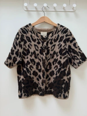Leopardmönstrad stickad kortärmad tröja - Säljer en supermjuk stickad tröja/kofta med leopardmönster i beige och svart. 🤎🩶🖤                        Tröjan har korta ärmar, knappar längst med och en  rund halsringning . 