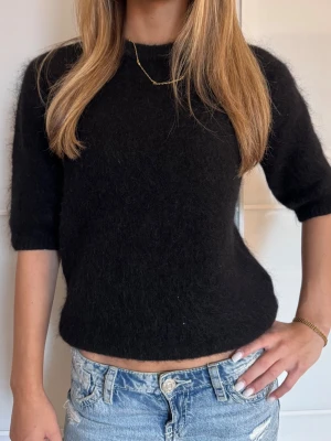 Svart stickad tröja med kort ärm - Mysig svart stickad tröja med korta ärmar och rund hals. Tröjan har en lätt fluffig yta och avslappnad passform, perfekt att styla till jeans eller kjol. Materialet ser ut att vara en mjuk ull- eller mohairblandning.