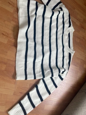 Vit stickad tröja med marinblå ränder - stickad tröja i vitt med marinblå  ränder. Tröjan har rund halsringning och ribbstickade muddar vid ärmslut och nederkant. Den är lite cropped och oversized. Inga tecken på användning så väldigt bra skick.