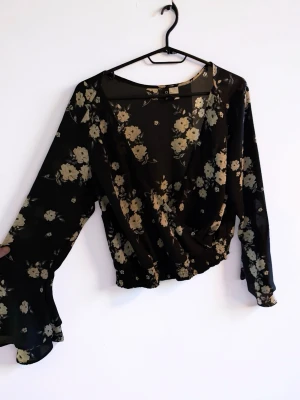 Svart blommig blus - Snygg svart omlottblus med beige blommönster och transparenta, vida långärmar. Blusen har djup v-ringning och resår i midjan för en smickrande passform. Perfekt att styla med jeans eller kjol för en trendig look.