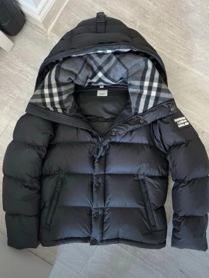 Svart pufferjacka med rutigt foder - Snygg svart pufferjacka med stor huva och rutigt grått/vitt foder. Jackan har dragkedja och knappar framtill, två sidofickor med dragkedja och en patch på ärmen. Perfekt för kalla vinterdagar och riktigt varm. I storlek står de xl men de passar l också