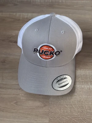 Grå och vit Pucko keps snapback - Snygg keps i grått och vitt med broderad Pucko-logga framtill. Klassisk snapback-modell med böjd skärm och mesh baktill för extra ventilation. Justerbar storlek med plastspänne. Perfekt för dig som gillar retrovibbar och streetwear.