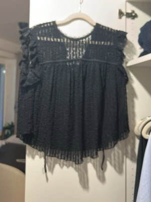 Svart spetsblus med volangdetaljer från NELLY - Säljer en svart blus i transparent spets med volangdetaljer på axlarna och knytband fram. Blusen har korta ärmar och ett luftigt, avslappnat fall. Perfekt för dig som gillar en romantisk och trendig stil.