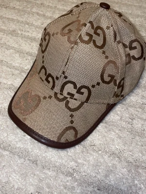 Beige keps med GG-mönster - Snygg beige keps med brunt GG-mönster och mörkbruna detaljer längs skärmen och sömmarna. Klassisk böjd skärm och justerbar passform. Materialet ser ut att vara canvas med läderliknande kant. Perfekt för dig som gillar streetwear och vill sticka ut.