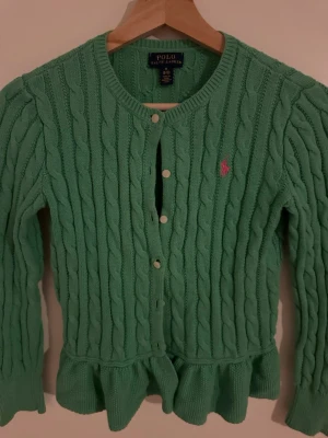 Polo Ralph Lauren/ barn  - Grön kabelstickad kofta från Polo Ralph Lauren med rund halsringning, volangkant nedtill och rosa broderad logga på bröstet. för barn/ flickor 