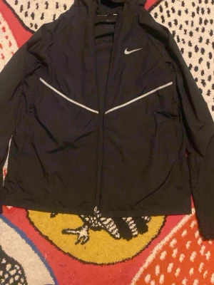 Svart löparjacka Nike Miler XL - Svart löparjacka från Nike Miler med hel dragkedja och reflekterande detaljer över bröstet. Jackan har hög krage och är tillverkad i lätt, funktionellt material som passar perfekt för löpning och träning utomhus.